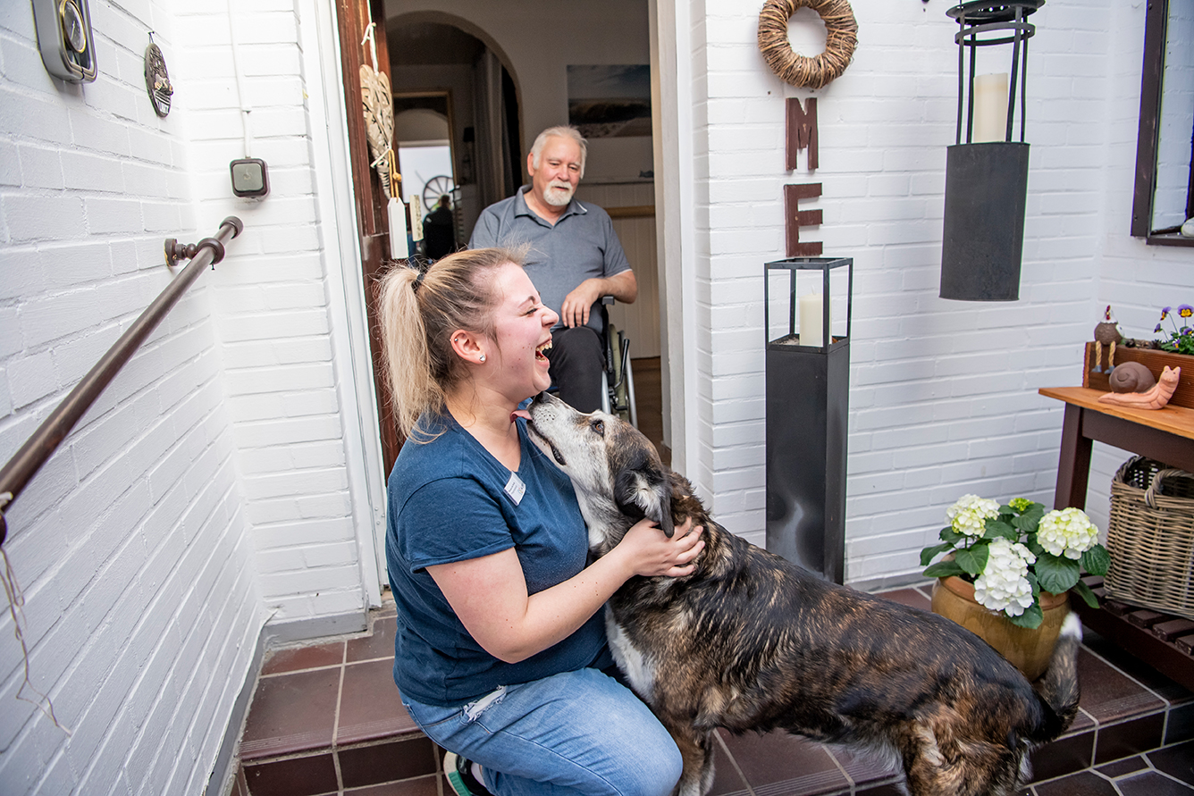 Weibliche Pflegefachkraft begrüßt lachend einen Hund vor der Haustür eines Klienten in Lehrte Sievershausen