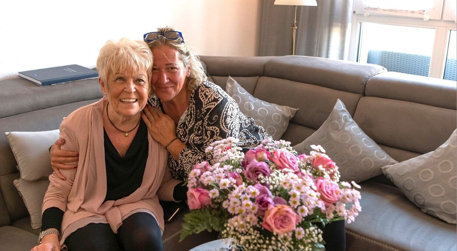 Pflegekraft umarmt lachende Seniorin auf Couch mit Blumen beim ambulanten Pflegedienst Stadtpark Hilden